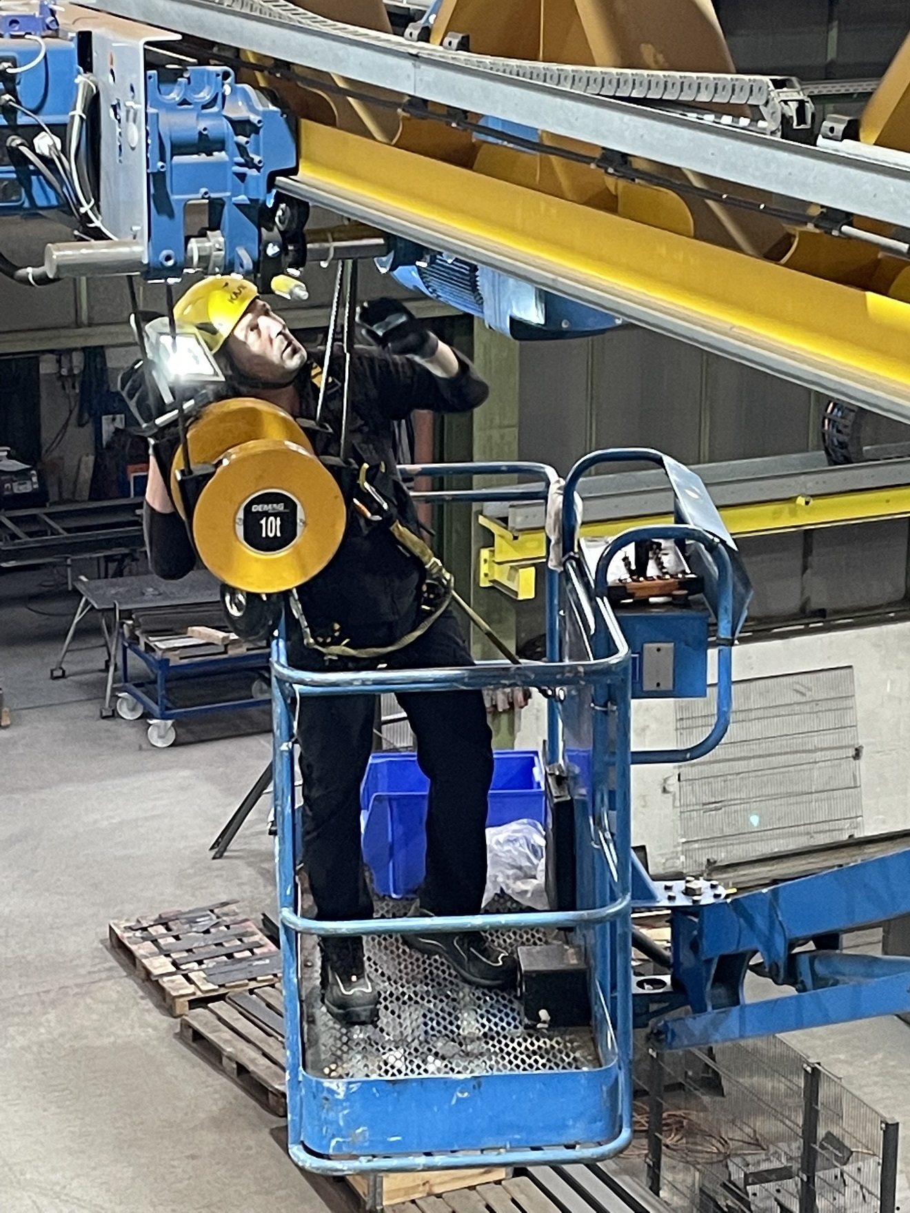 Crane.tool Kransachkundiger Service Mitarbeiter, auf einer Gelenkarbeitsbühne, bei der Prüfung eines DEMAG Seilzuges DMR an einem DEMAG Laufkran EVKE.